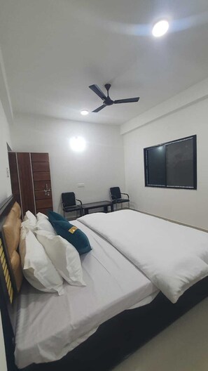 A hotel room with a bed, a desk, a chair, a wardrobe, and a ceiling fan. - US RESORT (Chainpur)