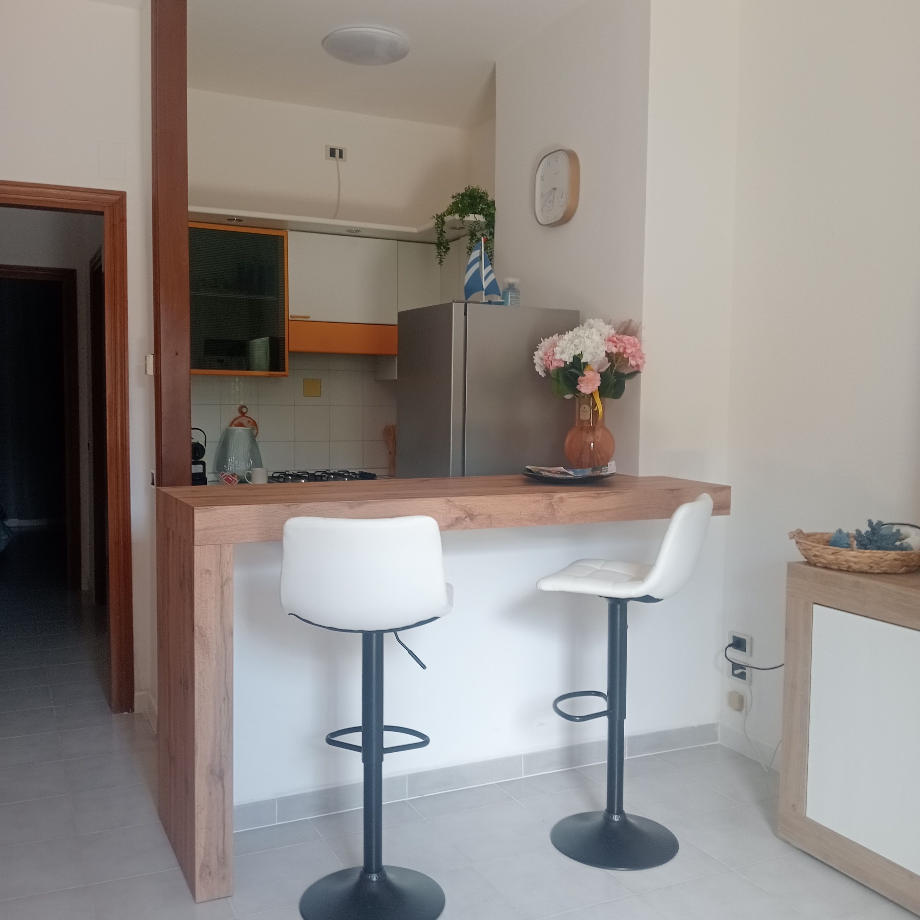 A modern kitchen with a wooden island, two bar stools, a refrigerator, and a clock on the wall.