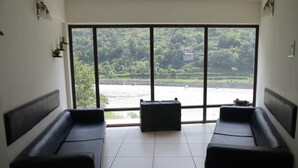 Una habitación con dos sofás negros, un otoman negro y un amplio ventanal con vistas a un río y zonas verdes.