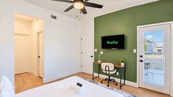 A hotel room with a bed, a desk with a chair, a ceiling fan, a TV mounted on the wall, and a door with a window.