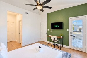 A hotel room with a bed, a desk with a chair, a ceiling fan, a TV mounted on the wall, and a door with a window. - Near TPC & Shops | Pool, Gym, Balcony & Parking (Scottsdale)