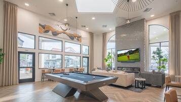 A modern living room with a pool table, comfortable seating, a large TV, and large windows offering a view of the outside.
