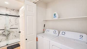 A laundry room with two white washing machines, a shelf with a detergent bottle, and a door leading to a bathroom with a glass shower.