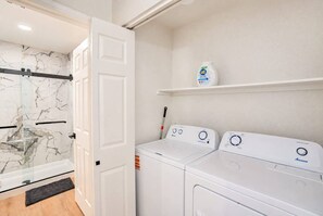 A laundry room with two white washing machines, a shelf with a detergent bottle, and a door leading to a bathroom with a glass shower. - Near TPC & Shops | Pool, Gym, Balcony & Parking (Scottsdale)