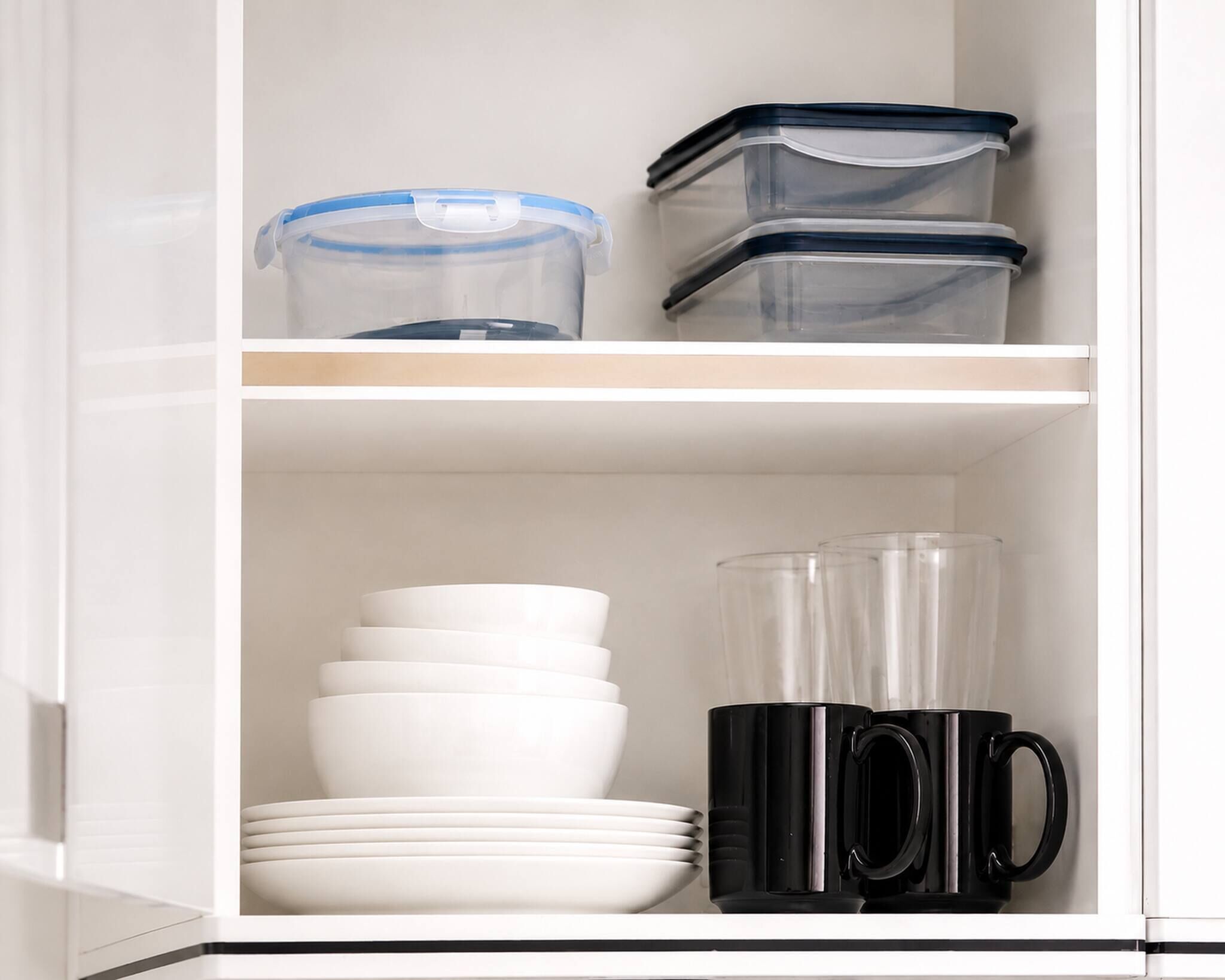 A kitchen cabinet with storage containers, white plates, and black mugs.