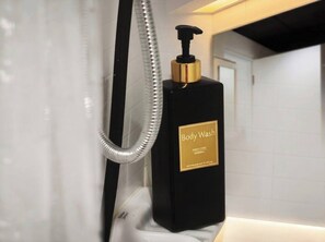 A black bottle of Body Wash with a gold label, a silver shower hose, and a white tiled wall. - S&G Beachfront Serviced 1-br, Step to Tops and 7-11 (Pattaya)