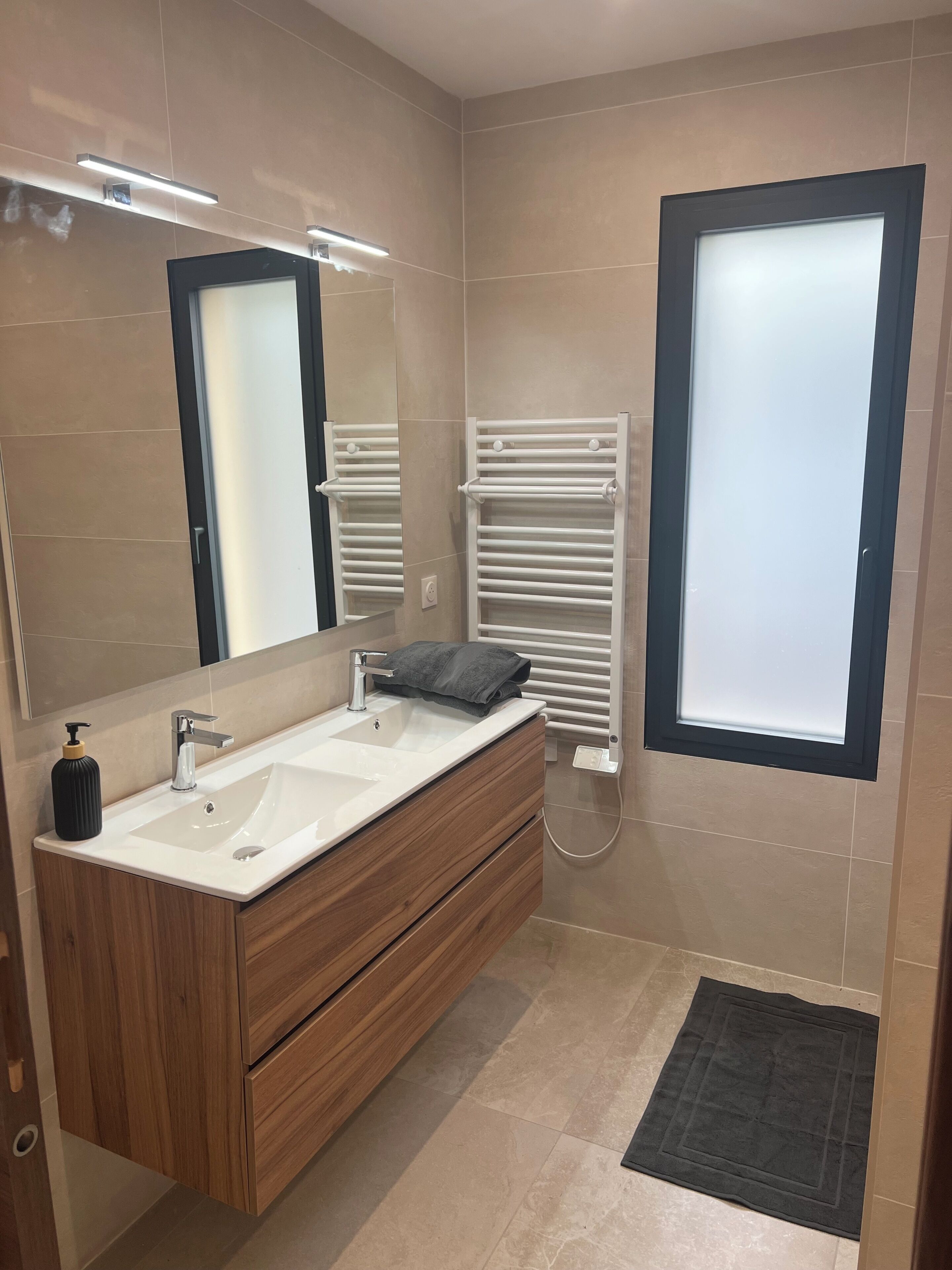 A modern bathroom with a wooden vanity, a large mirror, a white towel rack, and a window with frosted glass.
