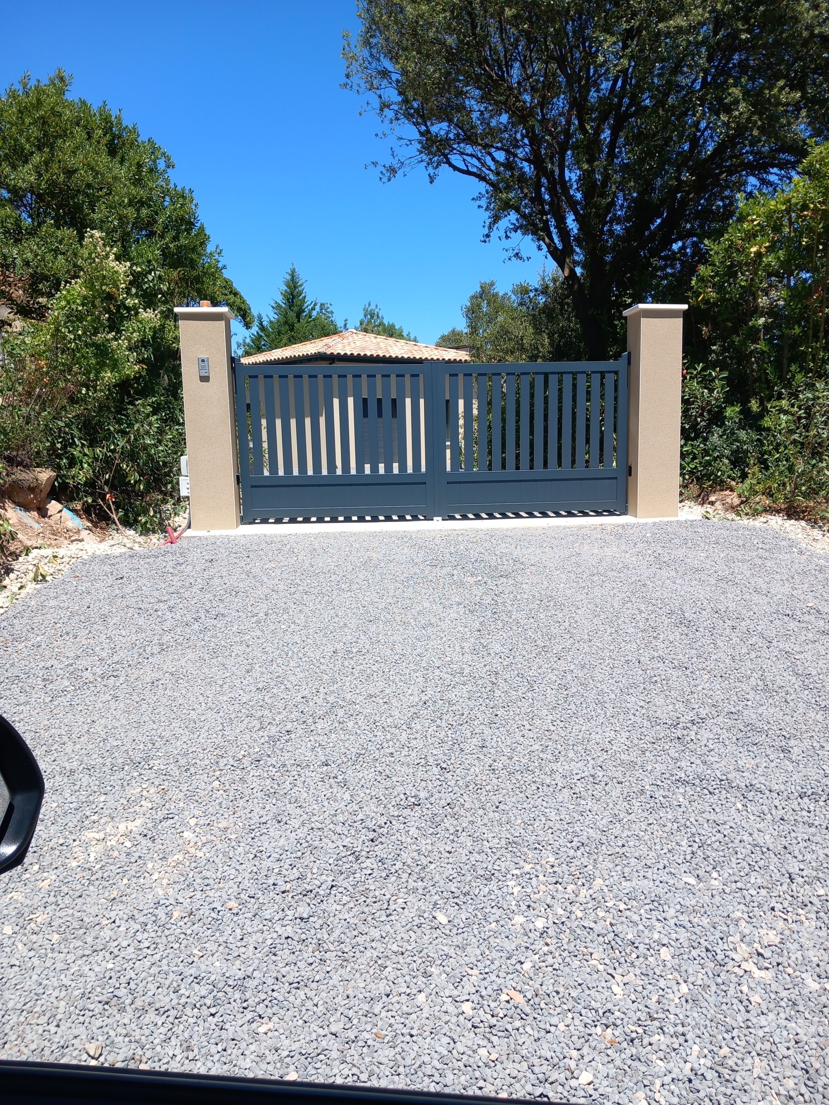 A gated driveway with a gravel surface, flanked by beige pillars, leading to a building with a tiled roof.