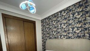A bedroom with a bed, a wooden wardrobe, a floral wallpaper, and a ceiling light fixture. - luxuary 3-bedroom apartment with AC in adorable Mohammédia (Mohammédia)