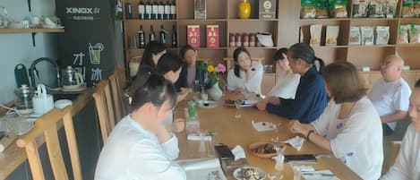 A group of people seated around a wooden table in a room with shelves stocked with bottles and packaged goods.