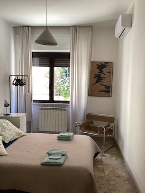 Ein Schlafzimmer mit einem Bett, einem Heizkörper, einem Fenster mit Vorhängen, einem Stuhl und einer Wand-Klimaanlage.