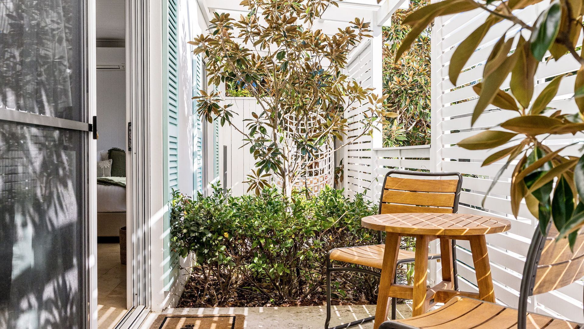 A small patio with a wooden chair and table, surrounded by plants and a white fence.