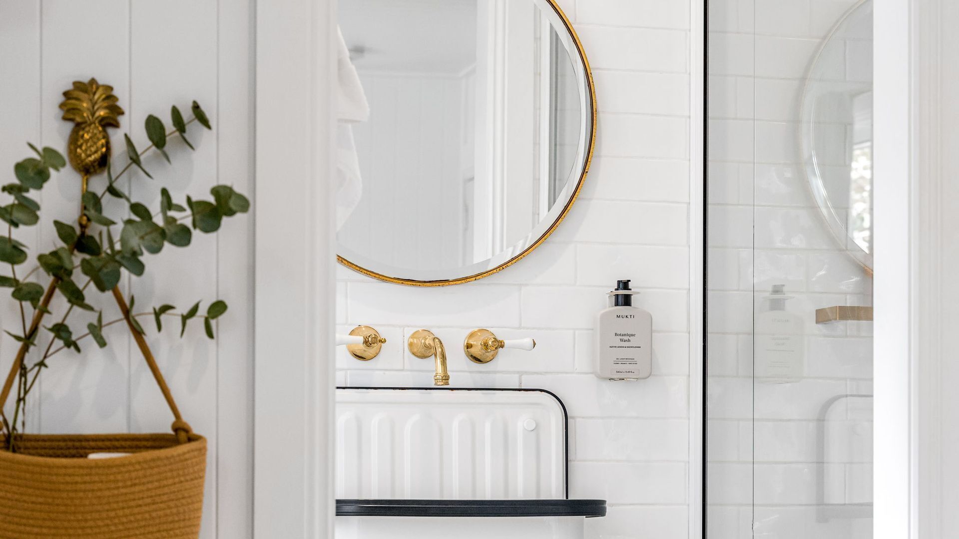 Un bagno con parete in piastrelle bianche, uno specchio con cornice dorata, un rubinetto dorato, una bottiglia di MUKTI Botanical Wash bianca e una pianta in un cesto di vimini.