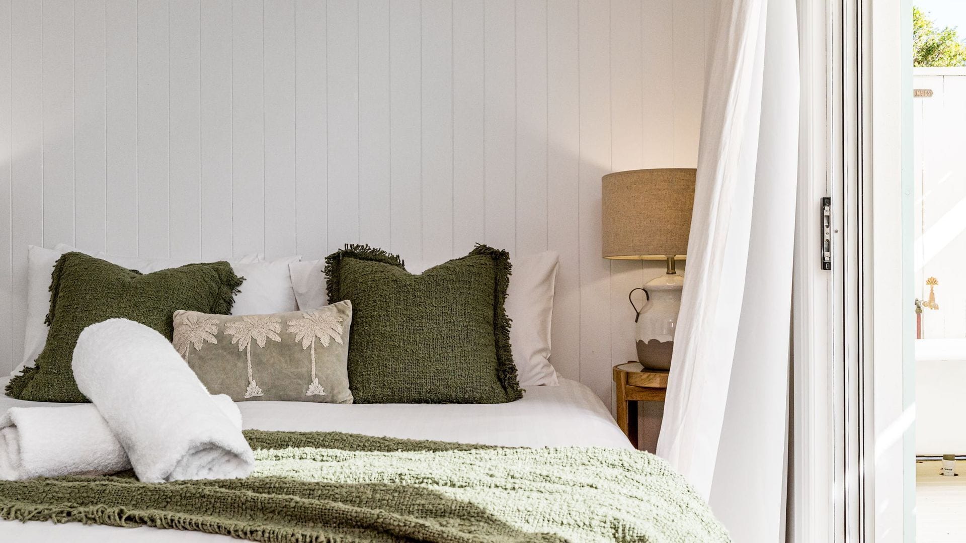 A neatly made bed with a green blanket and pillows, a bedside lamp, and a view of the outdoors through a window.