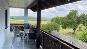 Un balcón techado con vistas a un paisaje verde, una mesa con sillas y una barandilla de madera.