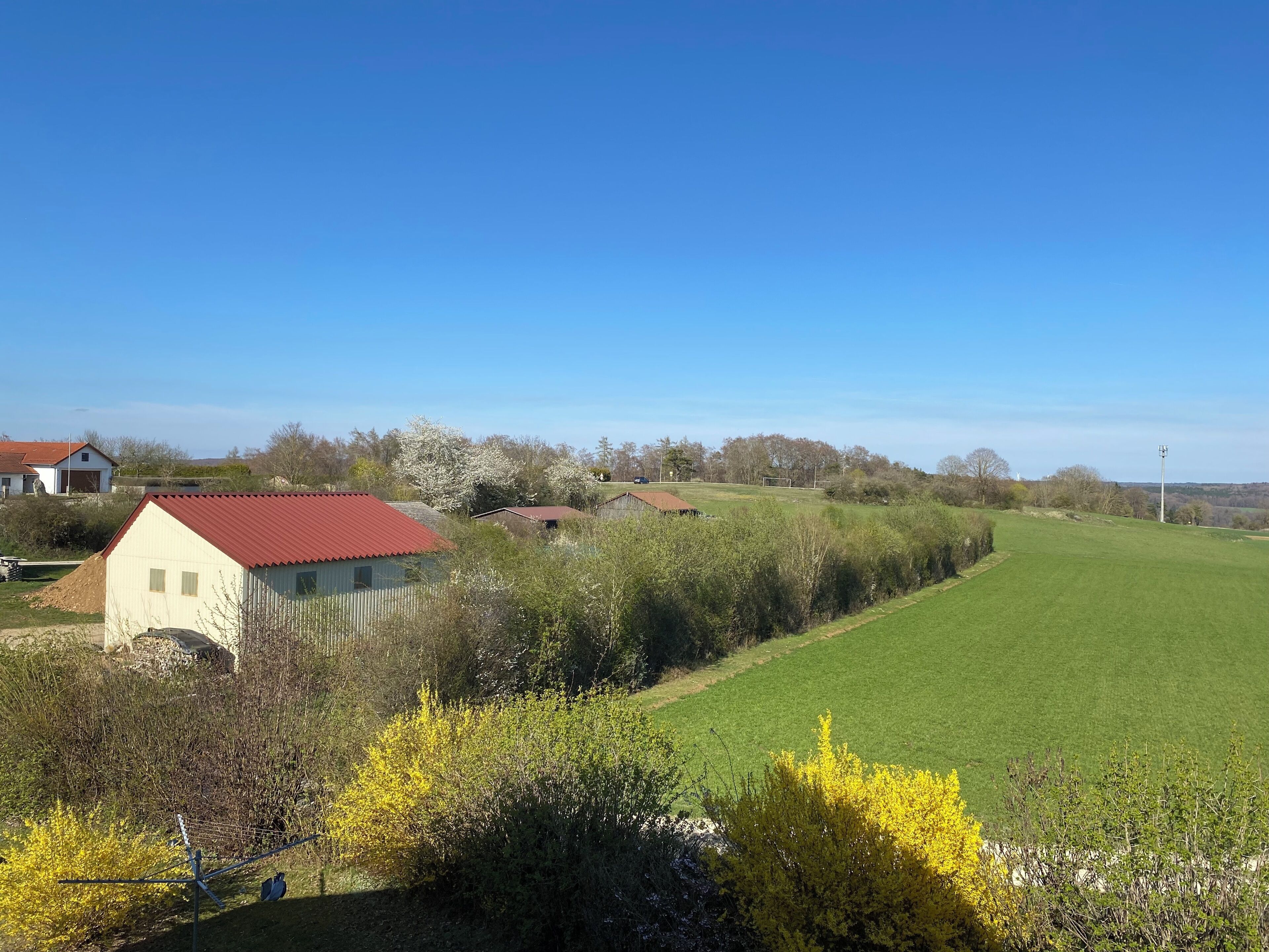 Ländliche Landschaft mit einem grünen Feld, einem kleinen Gebäude mit rotem Dach und blauem Himmel.