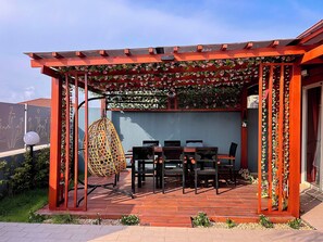 Ein überdachter Außenbereich mit Tisch und Stühlen, umgeben von einer roten Metallkonstruktion und Pflanzen.