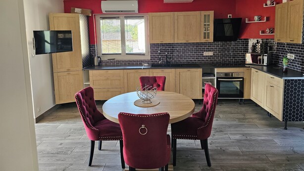 A modern kitchen with wooden cabinets, a round dining table with red chairs, a built-in oven, and a black tiled backsplash. - Under the stars in the countryside (Paray-le-Frésil)
