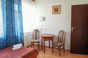 Una habitación con cama, dos sillas, una mesita, una puerta y un interruptor de luz en la pared.
