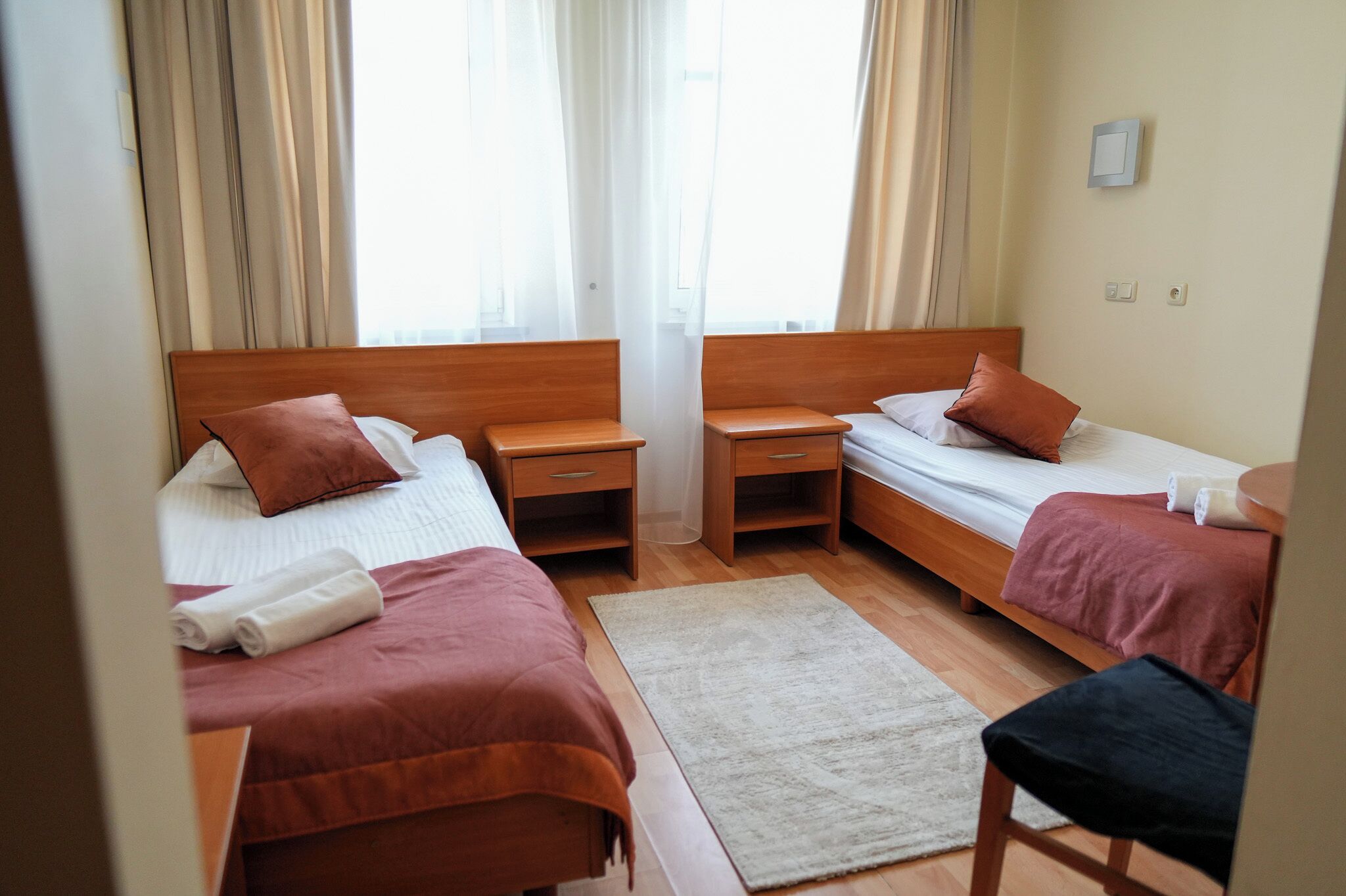 Una camera d'albergo con due letti singoli, una testiera in legno, un comodino con una lampada, un tappeto bianco e una sedia.