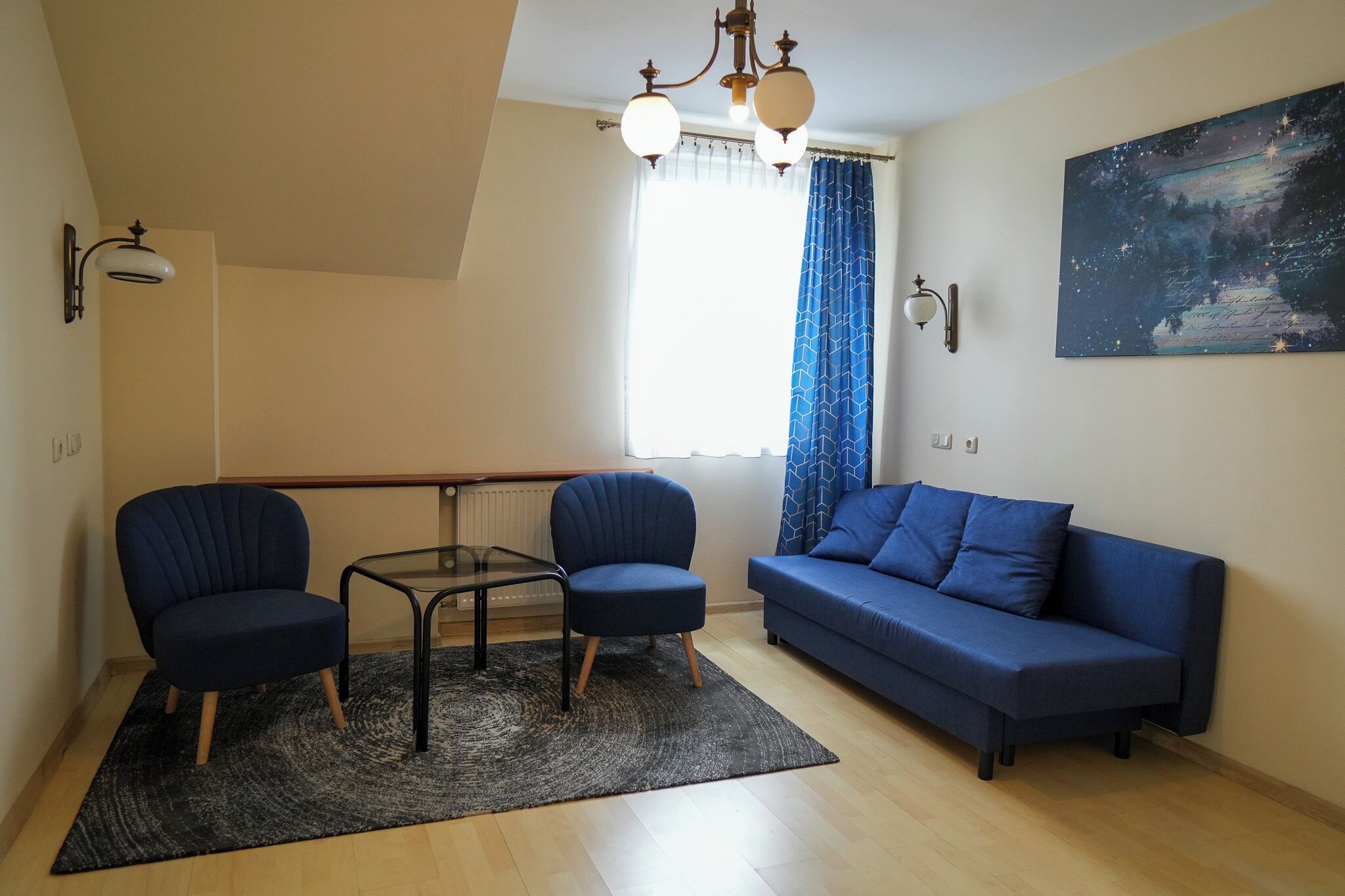 Uma sala de estar moderna com um sofá azul, duas poltronas, uma mesa de centro de vidro, um lustre e um quadro na parede.