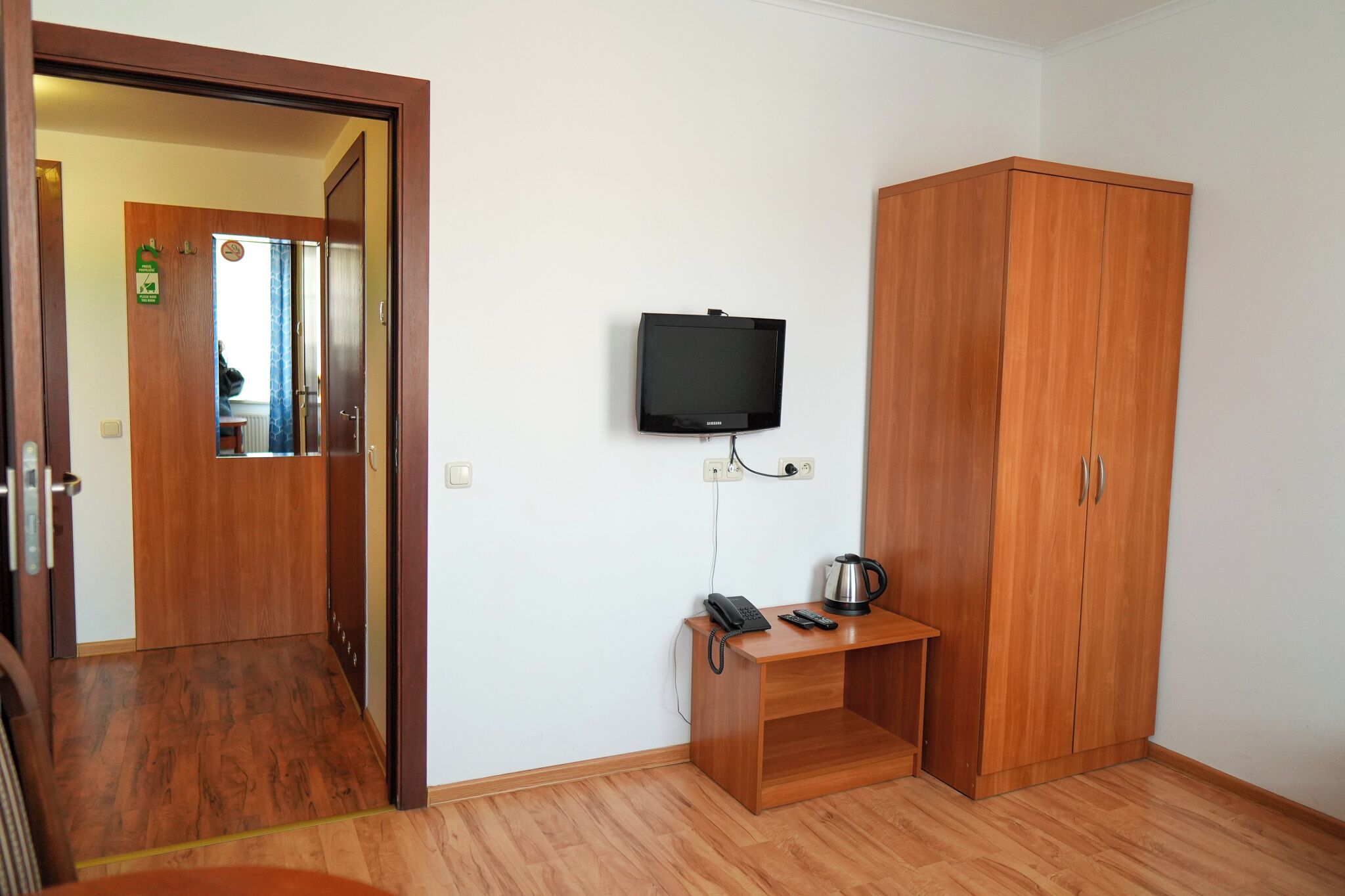 Una habitación con un armario de madera, un escritorio pequeño con teléfono y hervidor, y un televisor fijado a la pared.
