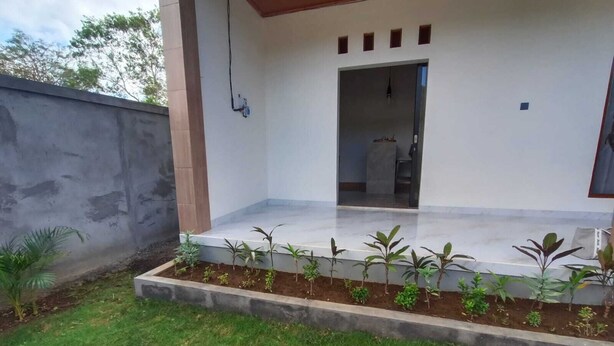 A covered outdoor area with a concrete floor, a small garden with young plants, and a doorway leading to an interior space.