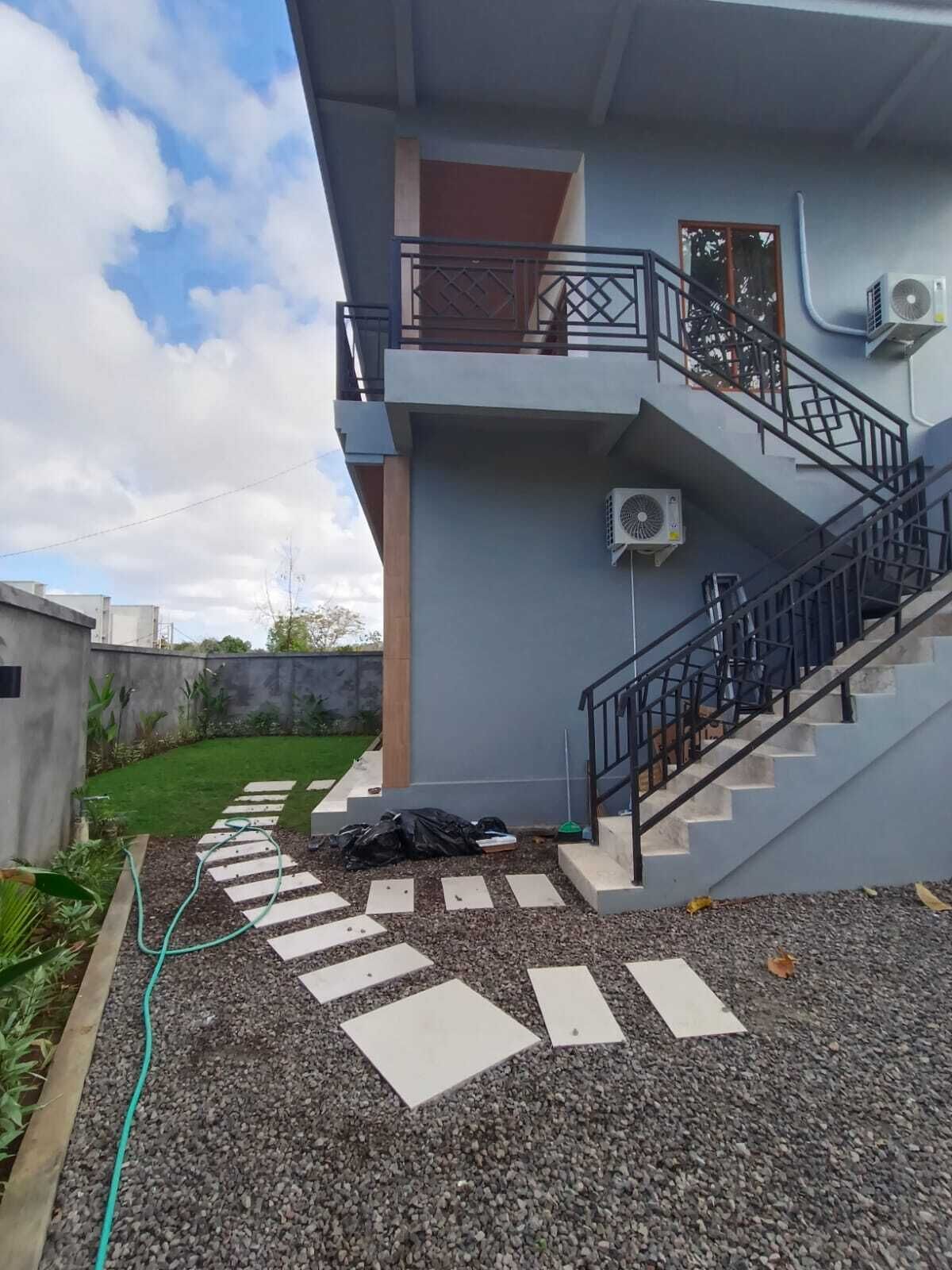 Un immeuble résidentiel doté d’un escalier, de climatiseurs et d’un espace jardin avec un tuyau d’arrosage et des pas japonais.