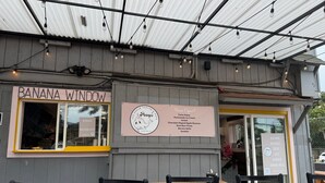 Un restaurante con un letrero que dice "BANANA WINDOW" y otro que indica "¡Abierto ya!” con una lista de platillos.