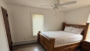 Ein Schlafzimmer mit einem Holzbett, Deckenventilator, einem Fenster mit Jalousien und Holzdielenboden.