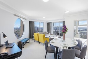 Ein moderner, gut beleuchteter Raum mit einem Esstisch für vier Personen, einem Sofa, einem Laptop auf einem Schreibtisch und großen Fenstern mit Blick auf die Stadt.