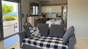 A modern living room with a sofa and a dining table, a kitchen with cabinets and appliances, and a view of the sea through a glass door. - Beach View House with Private Garden (ΧΑΛΚΙΔΑ)