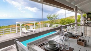 Ein Essbereich am Pool mit Blick auf das Meer, weiße Liegestühle und ein Tisch für zwei Personen.