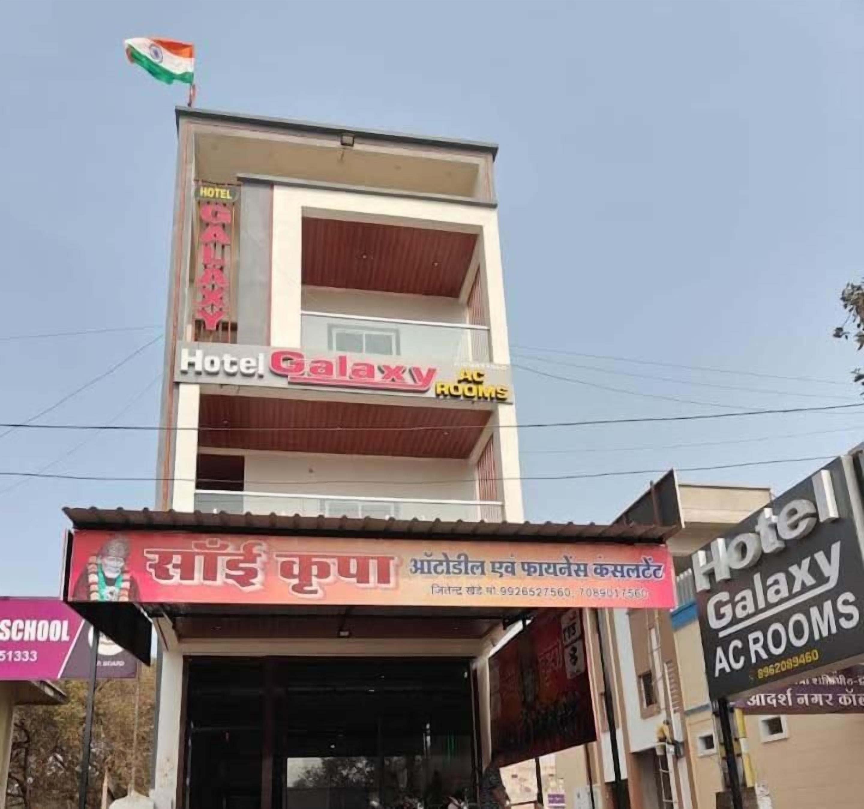 A two-story building with a sign for 'Hotel Galaxy AC Rooms' and another sign for 'Sain Kupa' with contact information.