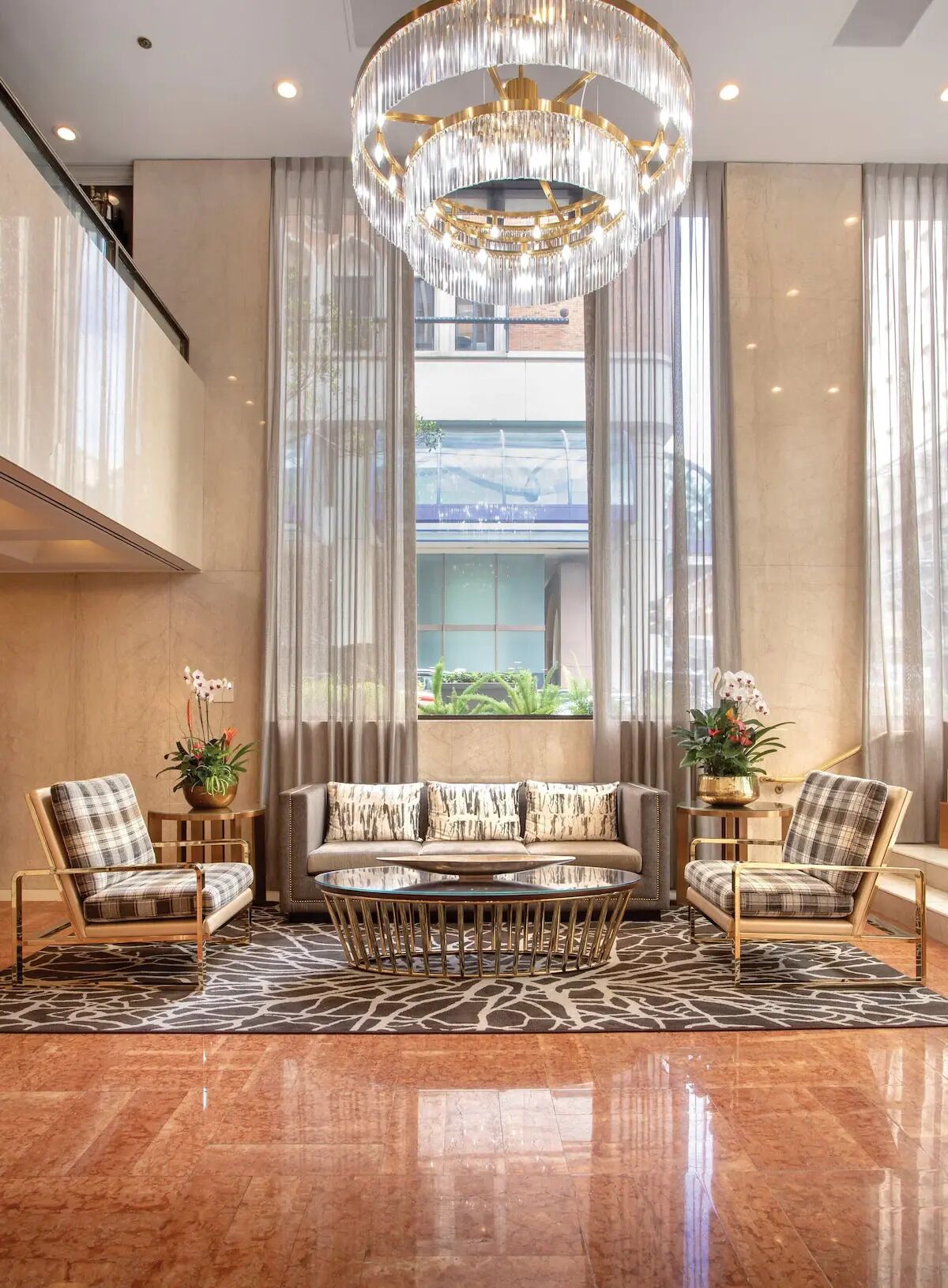 A modern lobby with a chandelier, a sofa, two armchairs, a glass coffee table, and a view of greenery outside.