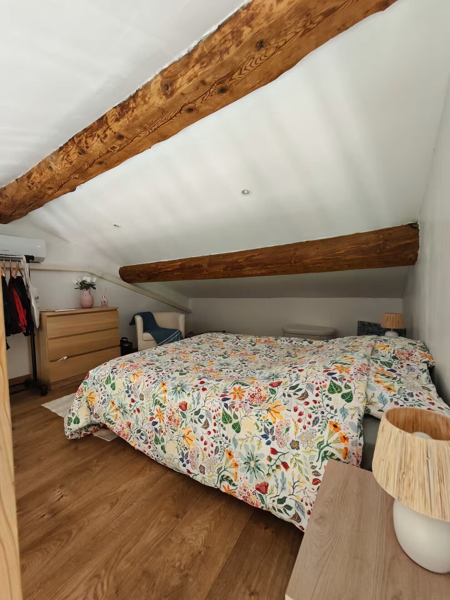 A bedroom with a sloped ceiling, wooden beams, a floral bedspread, a dresser, a chair, and a bedside table.