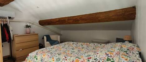 A bedroom with a sloped ceiling, wooden beams, a floral bedspread, a dresser, a chair, and a bedside table.