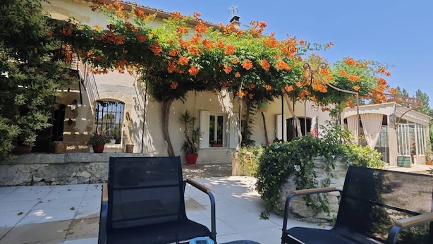 A patio with two black chairs, a table, and a building with a tiled roof and orange flowering vines. - One-bedroom apartment with a pool, in a Provençal farmhouse (Cavaillon)