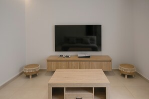 Ein modernes Wohnzimmer mit einem an der Wand befestigten Flachbildfernseher, einer Holztische mit Spielecontrollern, zwei gewebten Hockern und einem Couchtisch in der Mitte.