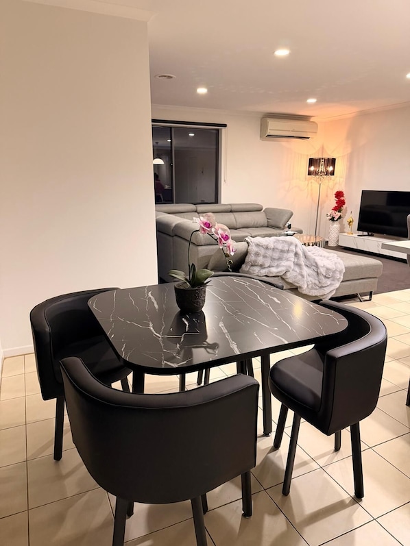 A modern living room with a dining area, including a black marble table and chairs, a grey sofa, and a flat-screen TV.