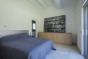 A bedroom with a bed, a wooden cabinet, a large framed cityscape picture, and a bathroom with a mirror and sink. - Villa L'Aparté Beauvallon | Pool, Movie Theater, Fitness Center & Golf (GRIMAUD)