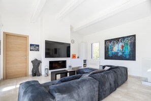 A modern living room with a large sectional sofa, a flat-screen TV, and artwork on the walls. - Villa L'Aparté Beauvallon | Pool, Movie Theater, Fitness Center & Golf (GRIMAUD)