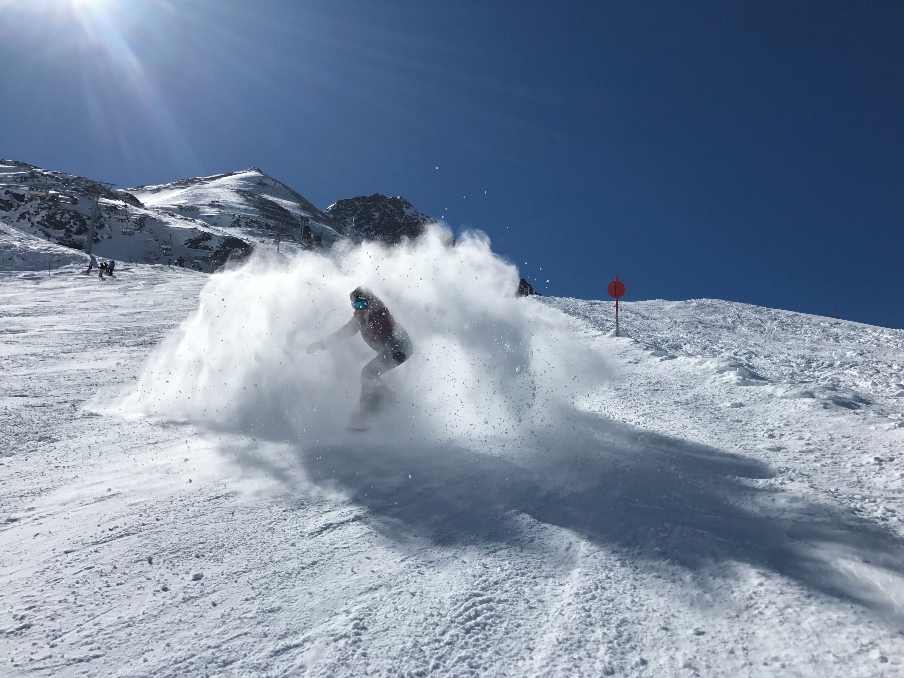 Ein verschneiter Berghang mit einer Person beim Snowboarden, eine Seilbahn im Hintergrund und einem klaren, blauen Himmel.