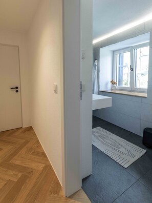 Une pièce avec un plancher en bois, une porte blanche, une fenêtre donnant sur un beau paysage, un lavabo blanc et un petit tapis.