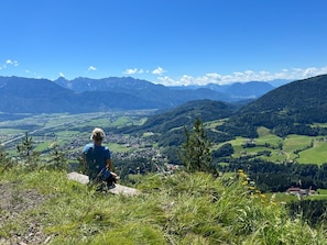 Une personne est assise sur une colline herbeuse, d’où elle a une vue sur une vallée, avec des montagnes en arrière-plan.