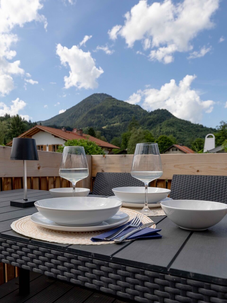 Un tavolo da pranzo all'aperto con piatti bianchi, due bicchieri di vino e posate, con vista su una montagna e un cielo sereno.