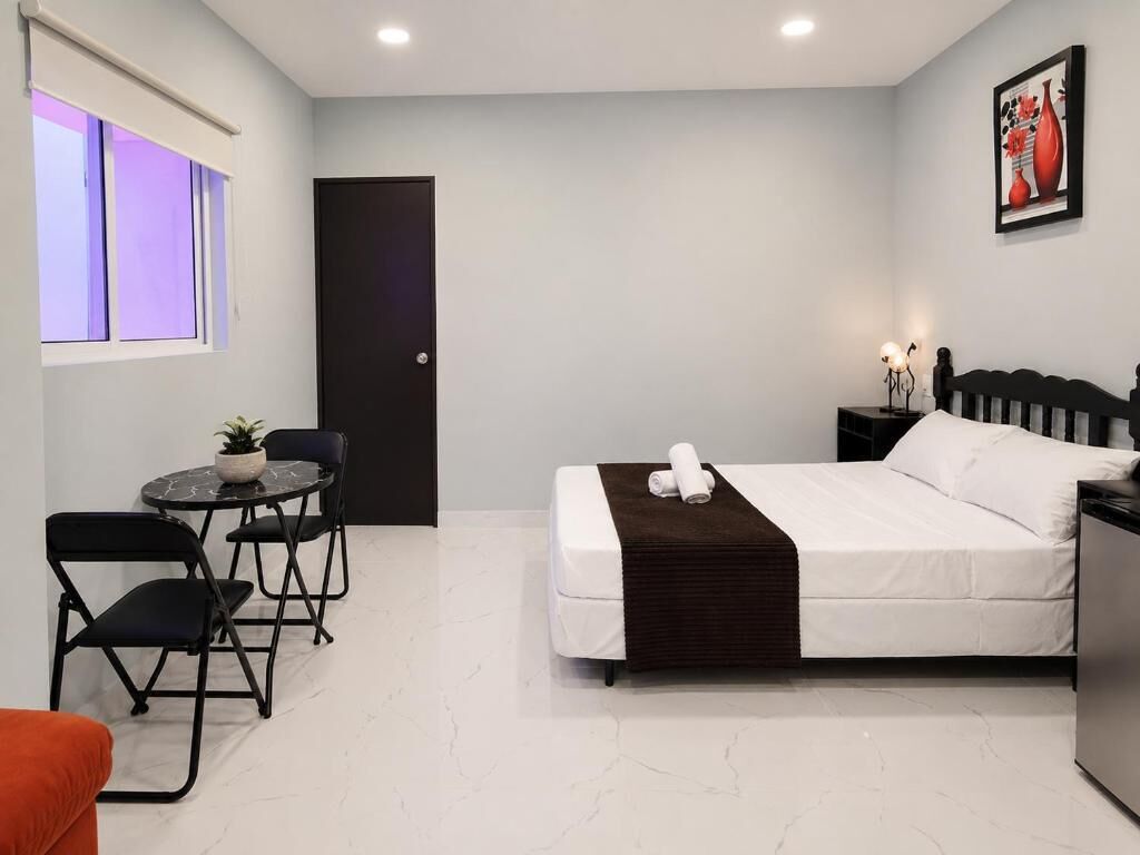 Une chambre d’hôtel moderne avec un lit, une petite table avec des chaises, une plante en pot, une porte et un tableau encadré au mur.