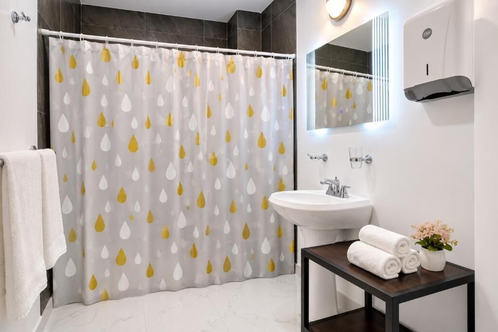Un baño con cortina de ducha, lavamanos, espejo y una mesita con toallas y flores.