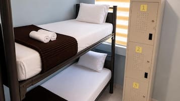 Una cama litera con ropa de cama blanca y cabeceros marrones, un armario metálico alto con compartimentos numerados, y una ventana con persianas.