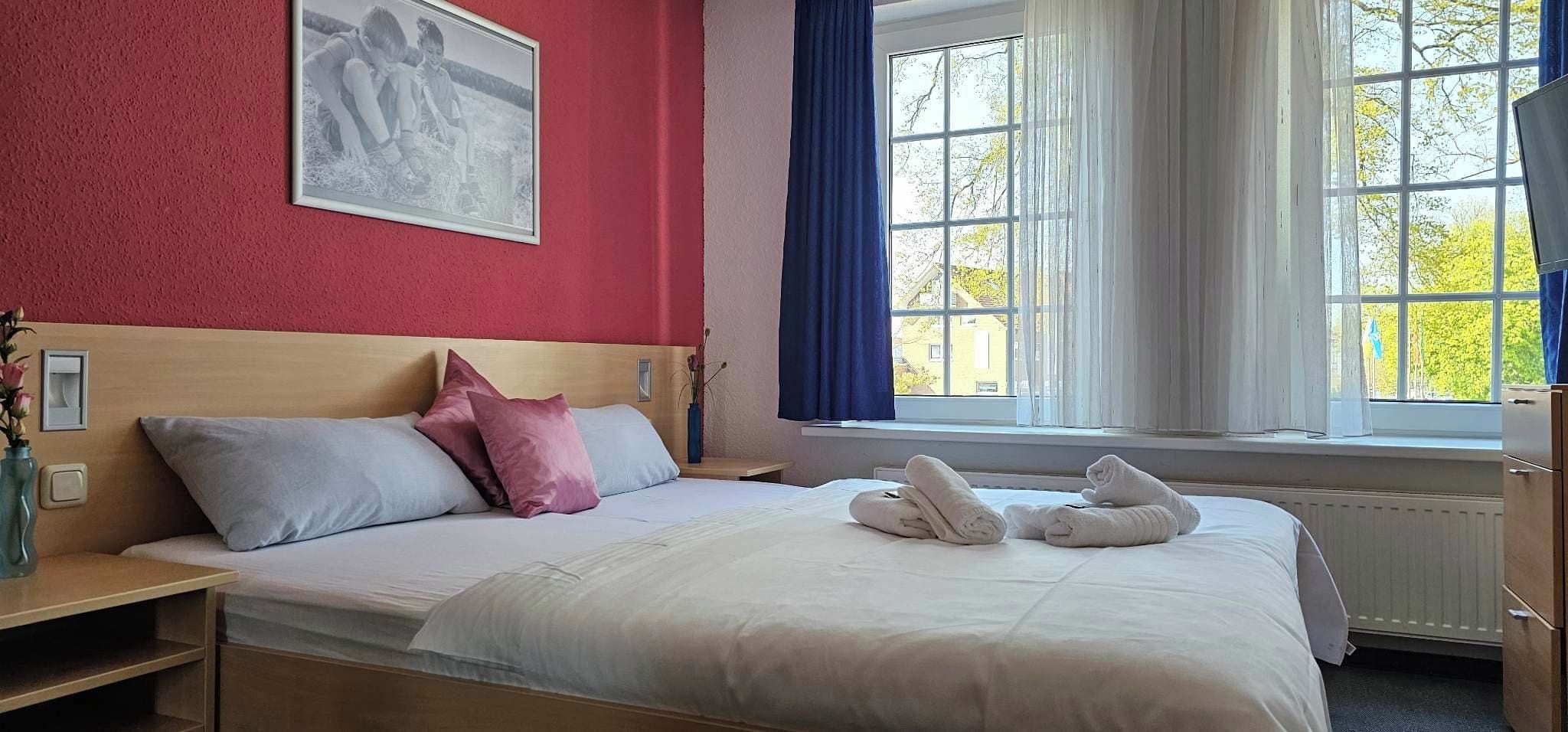 Habitación de hotel con cama, almohadas, mesita de noche con un florero, ventana con cortinas y un cuadro en la pared.